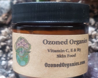 Puede incluir: Un frasco de vidrio marrón con tapa negra, etiquetado como "Ozoned Organics" con el texto "Vitamin C, E & B3 Skin Food" y el sitio web "OzonedOrganics.com".