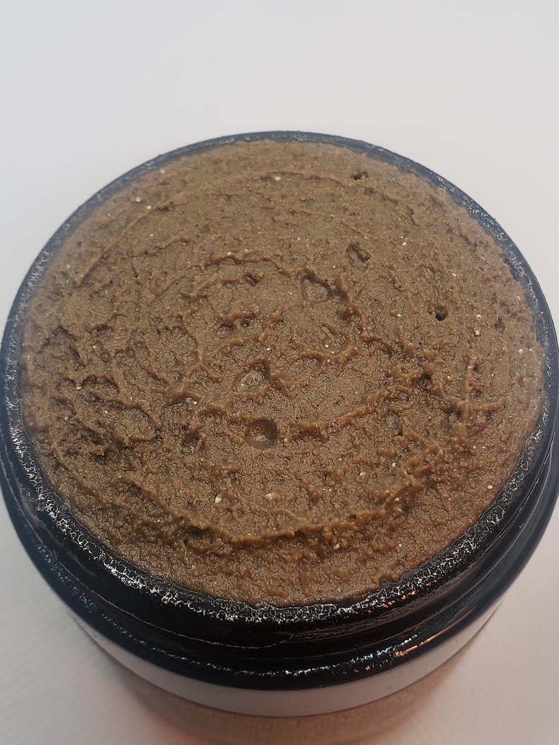 May include: A close-up of a brown, textured, creamy substance in a black jar. The substance appears to be a facial scrub or mask.