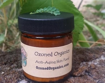 May include: A small, brown glass jar with a black lid, labeled "Ozoned Organics Anti-Aging Skin Food." A sprig of green leaves rests on top. The jar sits on a mossy surface, with green foliage in the background.