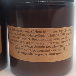 May include: A brown glass jar with a label listing the ingredients of a supplement. The ingredients include coconut oil, calcium bentonite clay, baking soda, xylitol, probiotic powder, theobromine, cacao powder, cinnamon, 72 trace mineral supplement, vitamin D3, vitamin K2, wheatgrass powder, distilled water, peppermint essential oil. The label also states that the product is all organic, vegan, and non-GMO.