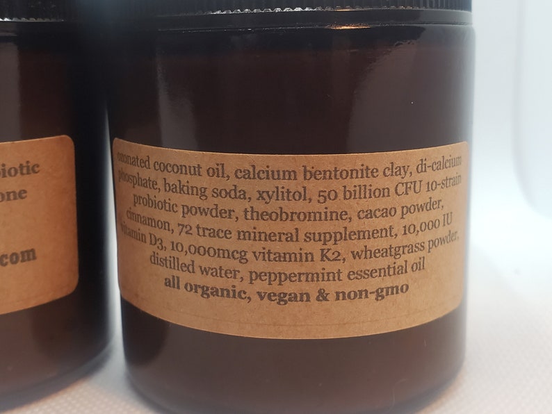 May include: A brown glass jar with a label listing the ingredients of a supplement. The ingredients include coconut oil, calcium bentonite clay, baking soda, xylitol, probiotic powder, theobromine, cacao powder, cinnamon, 72 trace mineral supplement, vitamin D3, vitamin K2, wheatgrass powder, distilled water, peppermint essential oil. The label also states that the product is all organic, vegan, and non-GMO.