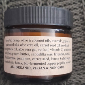 Op de afbeelding: Een bruine glazen pot met een zwarte deksel, gelabeld met een lijst van ingredi&euml;nten, waaronder hennep, olijf, kokos, avocado, jojoba, druivenpitolie, alo&euml; vera-olie, wortelzaadolie, rozenbottelolie, granaatappelolie, alo&euml; vera-gel, retinol, vitamine C, hyaluronzuur, hennepzaadboter, candelillawas, lavendel, neroli, wierook, geranium, wortelzaad, citroen, clary salie, etherische oli&euml;n, boorax, bio-gefermenteerd koperpeptidepoeder. Het etiket vermeldt ook "ALL ORGANIC, VEGAN & NON-GMO".