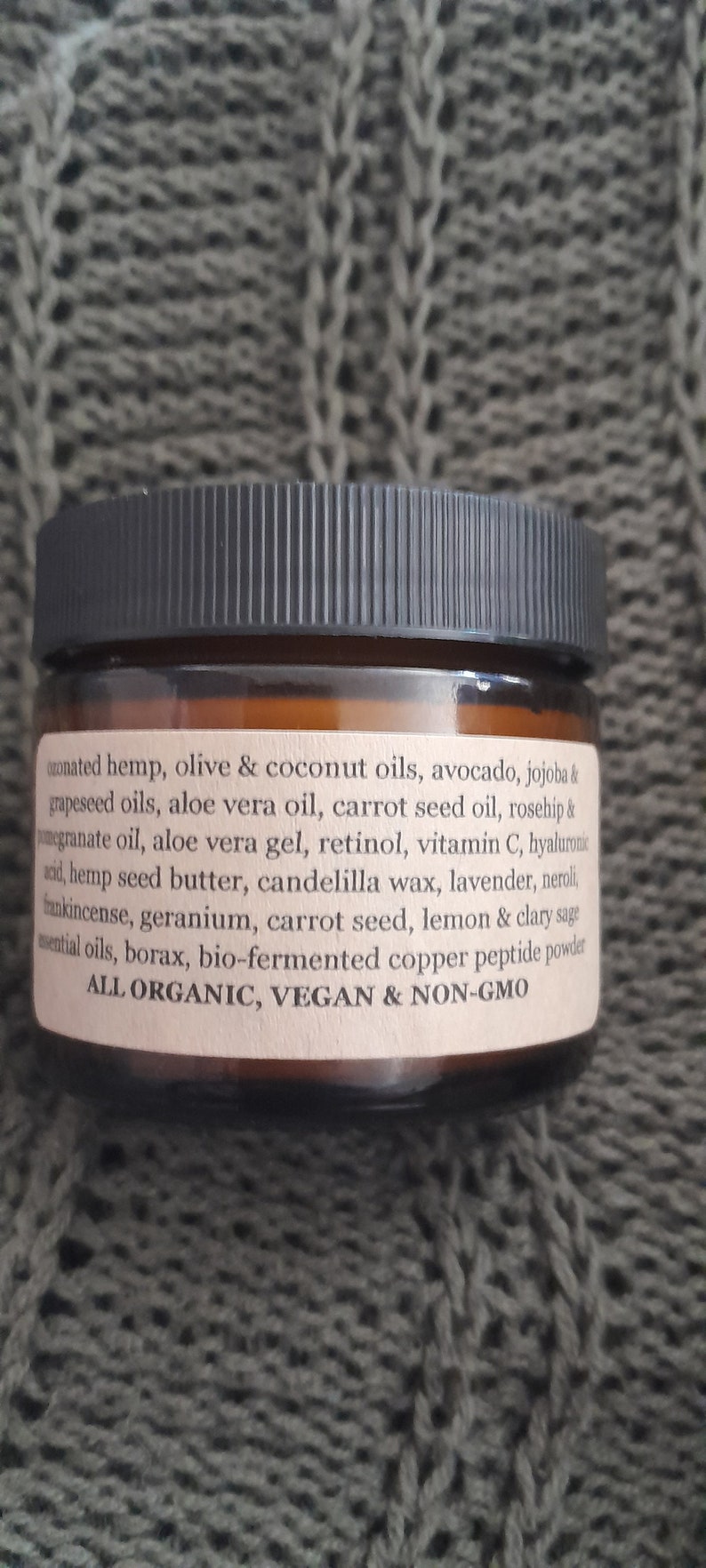 Op de afbeelding: Een bruine glazen pot met een zwarte deksel, gelabeld met een lijst van ingredi&euml;nten, waaronder hennep, olijf, kokos, avocado, jojoba, druivenpitolie, alo&euml; vera-olie, wortelzaadolie, rozenbottelolie, granaatappelolie, alo&euml; vera-gel, retinol, vitamine C, hyaluronzuur, hennepzaadboter, candelillawas, lavendel, neroli, wierook, geranium, wortelzaad, citroen, clary salie, etherische oli&euml;n, boorax, bio-gefermenteerd koperpeptidepoeder. Het etiket vermeldt ook "ALL ORGANIC, VEGAN & NON-GMO".