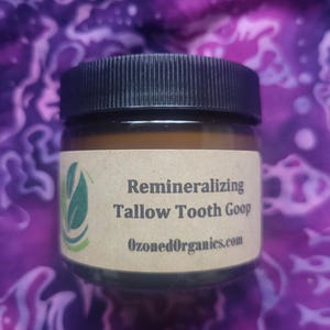 Peut inclure: Petit pot avec un couvercle noir et une étiquette beige. L'étiquette indique "Remineralizing Tallow Tooth Goop" avec le site Web "OzonedOrganics.com" en dessous. Un motif de feuille verte est sur le côté gauche de l'étiquette. Le fond est un motif tie-dye violet et blanc.