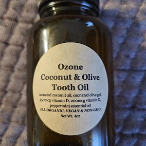 Könnte beinhalten: Eine braune Glasflasche mit einem weißen Etikett, auf dem "Ozone Coconut & Olive Tooth Oil" steht und die Zutaten aufgelistet sind: ozoniertes Kokosnussöl, ozoniertes Olivenöl-Gel, 250 mcg Vitamin D, 200 mcg Vitamin K, Pfefferminzöl. Das Etikett enthält auch die Angaben "All Organic, Vegan & Non-GMO" und "Net Wt. 4oz".