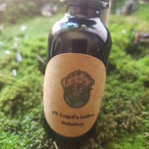 Op de afbeelding: Een klein donkerbruin glazen flesje met een zwarte dop en een bruin papieren etiket. Het etiket heeft een afbeelding van een wereldbol met een bloemdessin en de tekst "2% Lugol's Iodine Solution".