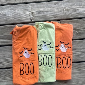 May include: Three t-shirts in orange, light green, and orange, each featuring a chest pocket embroidered with the word BOO in black lettering, accompanied by a white ghost and several small black bats stitched above the pocket opening.