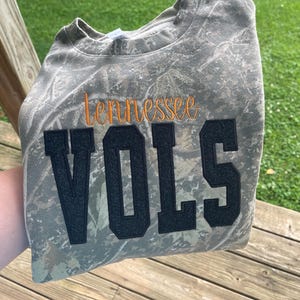 May include: A crewneck sweatshirt featuring a camouflage pattern in shades of brown, green, and tan. The word "tennessee" is embroidered in orange script above the word "VOLS," which is displayed in large, black, glittery block letters.