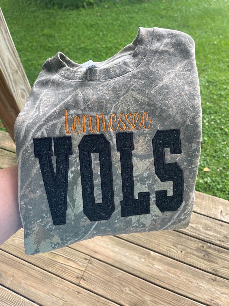 May include: A crewneck sweatshirt featuring a camouflage pattern in shades of brown, green, and tan. The word "tennessee" is embroidered in orange script above the word "VOLS," which is displayed in large, black, glittery block letters.