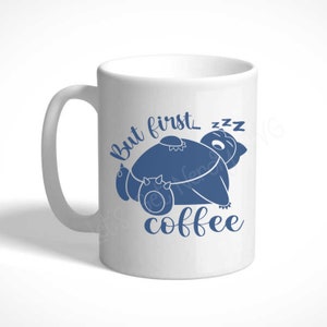 Może przedstawiać: Biały ceramiczny kubek do kawy z niebieskim nadrukiem śpiącego stworzenia z kresk&oacute;wek i tekstem "But first... coffee"