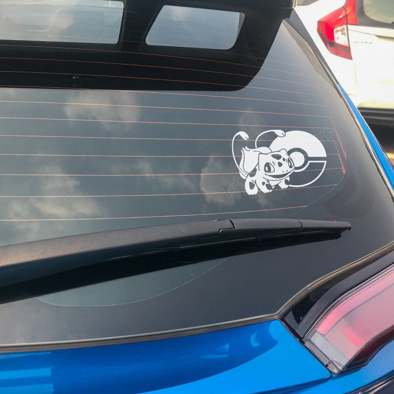 May include: A blue car with a white vinyl sticker of a Pokemon character on the back window. The sticker shows a white Pokemon character with a red spot on its back, holding a Pokeball.