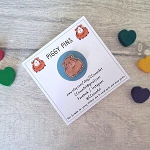 May include: A brown guinea pig pin on a blue background. The pin is part of a set of piggy pins. The card reads "Piggy Pins" and includes contact information for the shop.