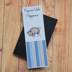 May include: A white bookmark with blue and brown stripes. The bookmark features a cartoon illustration of a small animal with the text "Popcorn With Happiness".
