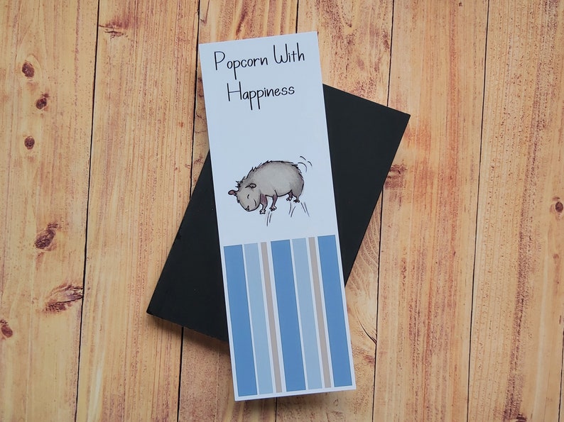May include: A white bookmark with blue and brown stripes. The bookmark features a cartoon illustration of a small animal with the text "Popcorn With Happiness".