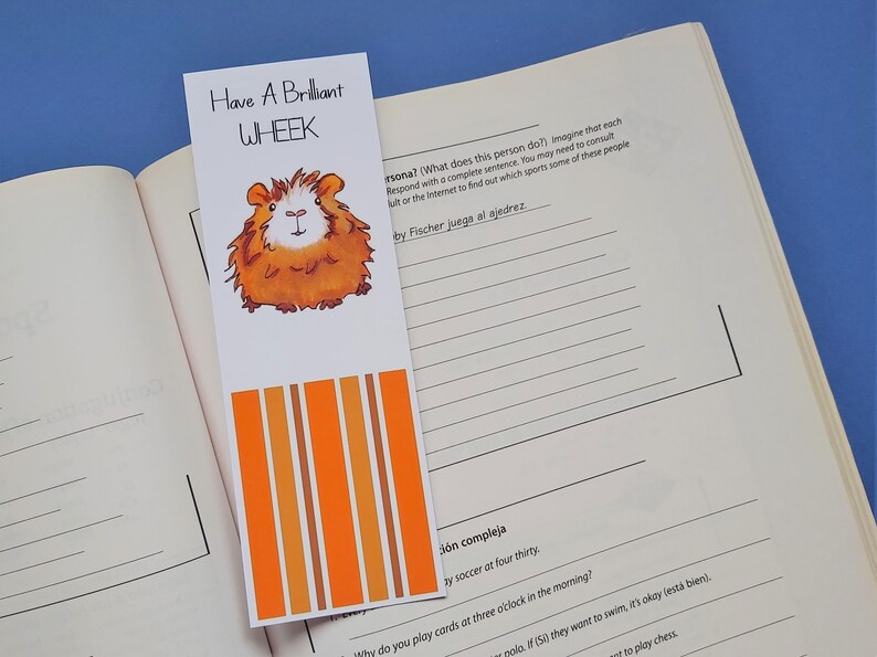 May include: A white bookmark with orange and brown stripes. The bookmark features a cartoon illustration of a brown and white guinea pig. The text "Have A Brilliant WHEEK" is printed on the bookmark.
