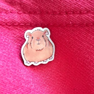 May include: A brown and white cartoon guinea pig sticker with a white outline. The sticker is on a pink fabric background.