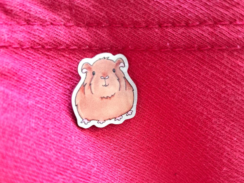May include: A brown and white cartoon guinea pig sticker with a white outline. The sticker is on a pink fabric background.