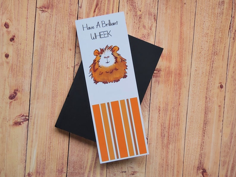May include: A white bookmark with orange and brown stripes. The bookmark features a cartoon illustration of a guinea pig and the text "Have A Brilliant WHEEK".