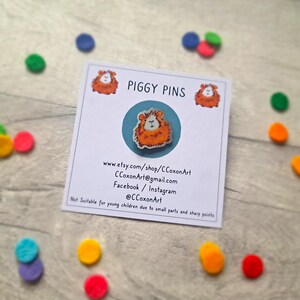 May include: A white card with the text "PIGGY PINS" printed in black. The card also includes a website address, email address, and social media handles. A small, orange and white guinea pig pin is displayed on the card.