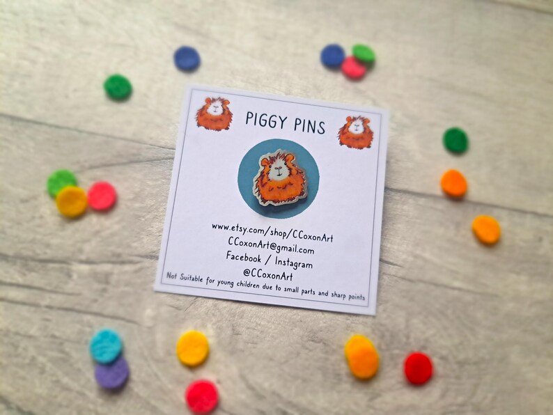 May include: A white card with the text "PIGGY PINS" printed in black. The card also includes a website address, email address, and social media handles. A small, orange and white guinea pig pin is displayed on the card.
