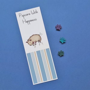 May include: A white bookmark with blue and beige stripes. The bookmark features a cartoon illustration of a brown dog with the text "Popcorn With Happiness" at the top.