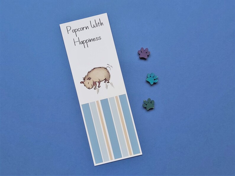 May include: A white bookmark with blue and beige stripes. The bookmark features a cartoon illustration of a brown dog with the text "Popcorn With Happiness" at the top.