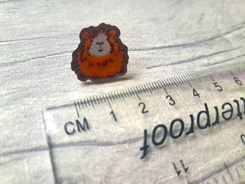 May include: A close-up of a small, orange and white guinea pig pin. The pin is about 1 cm in diameter and is made of metal.