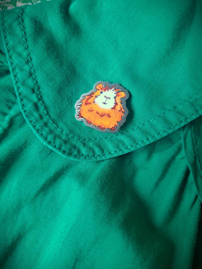 May include: A silver pin with a painted image of a brown and white guinea pig. The guinea pig is looking directly at the viewer.