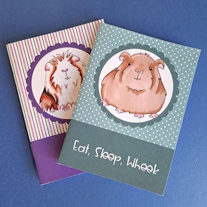 Peut inclure: Deux cartes de vœux avec des illustrations de cochons d'Inde. L'une a un fond rayé avec un cochon d'Inde dans un cadre festonné. L'autre a un fond à pois avec un cochon d'Inde et le texte "Eat, Sleep, Wheek".