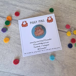 May include: A blue and white enamel pin featuring a cartoon guinea pig. The pin is on a white card with the text "PIGGY PINS" and contact information for the seller.