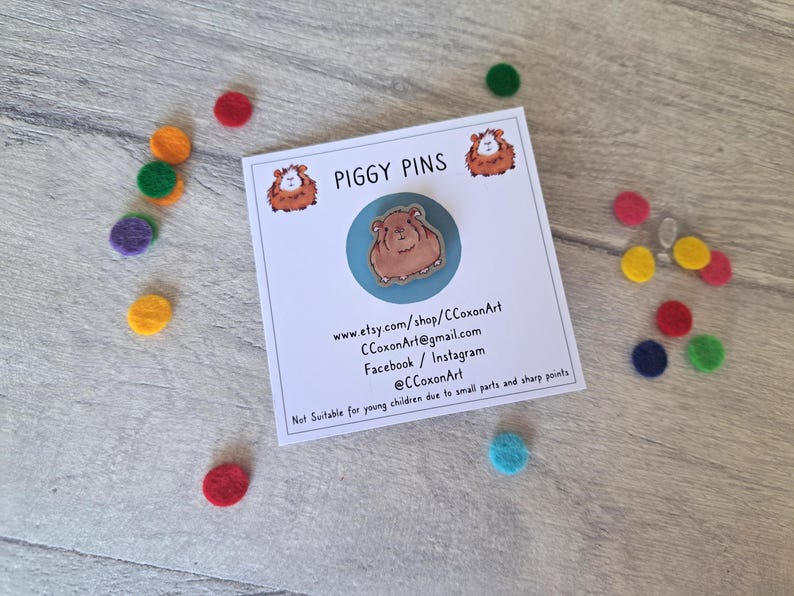 May include: A blue and white enamel pin featuring a cartoon guinea pig. The pin is on a white card with the text "PIGGY PINS" and contact information for the seller.