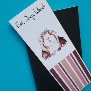 May include: A white bookmark with a cartoon guinea pig and the text "Eat, Sleep, Wheek". The bookmark has brown, pink, and beige stripes at the bottom.