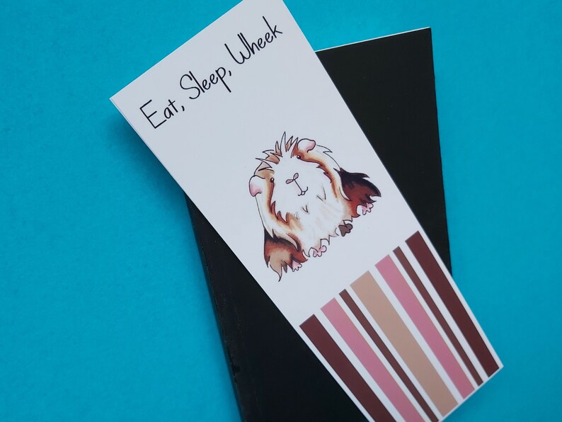 May include: A white bookmark with a cartoon guinea pig and the text "Eat, Sleep, Wheek". The bookmark has brown, pink, and beige stripes at the bottom.