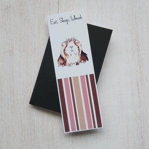 May include: A white bookmark with brown and pink stripes. The bookmark features a cartoon illustration of a guinea pig with the text "Eat, Sleep, Wheek".