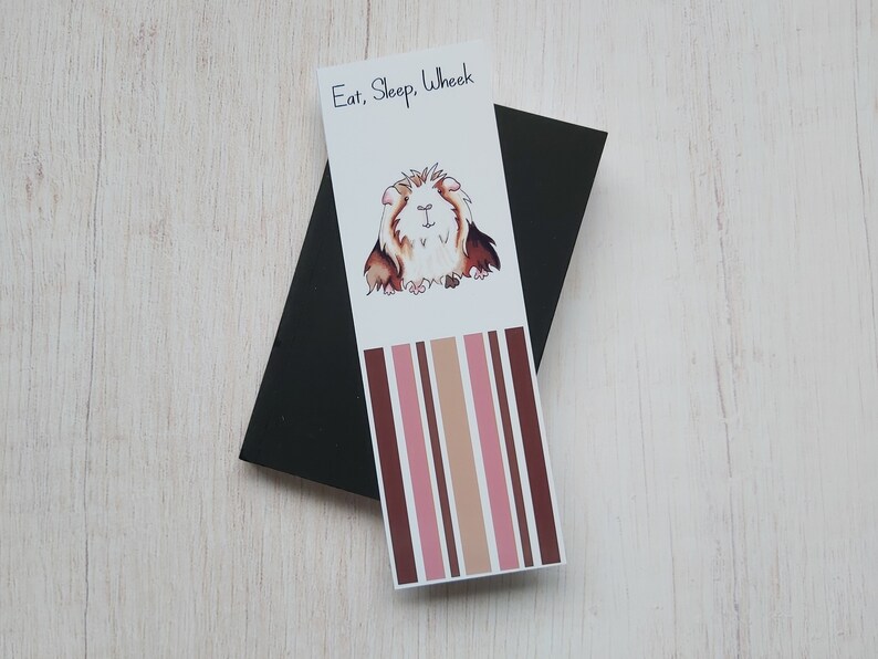 May include: A white bookmark with brown and pink stripes. The bookmark features a cartoon illustration of a guinea pig with the text "Eat, Sleep, Wheek".
