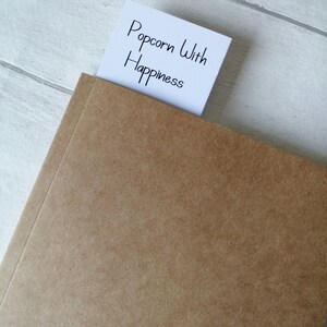 May include: A brown notebook with a white sticky note that says "Popcorn With Happiness" sticking out of the top.