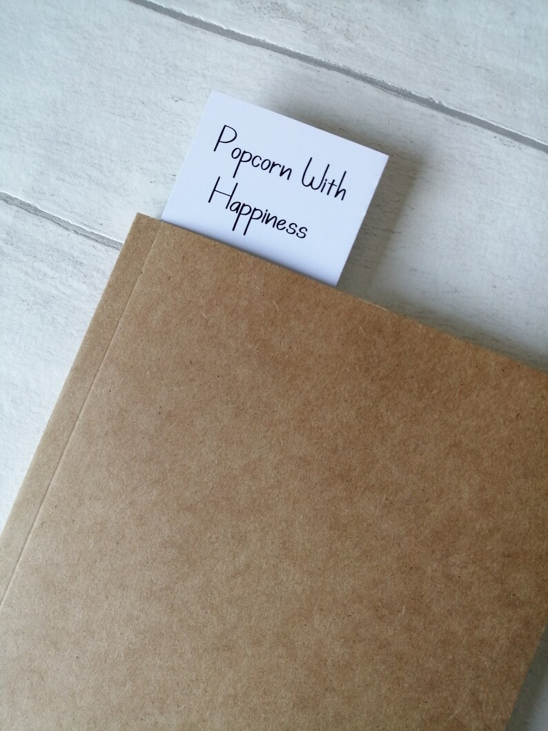 May include: A brown notebook with a white sticky note that says "Popcorn With Happiness" sticking out of the top.