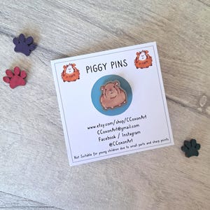 May include: A brown and white guinea pig pin on a blue background. The pin is part of a set of piggy pins. The card includes the shop name, website, and social media handles.