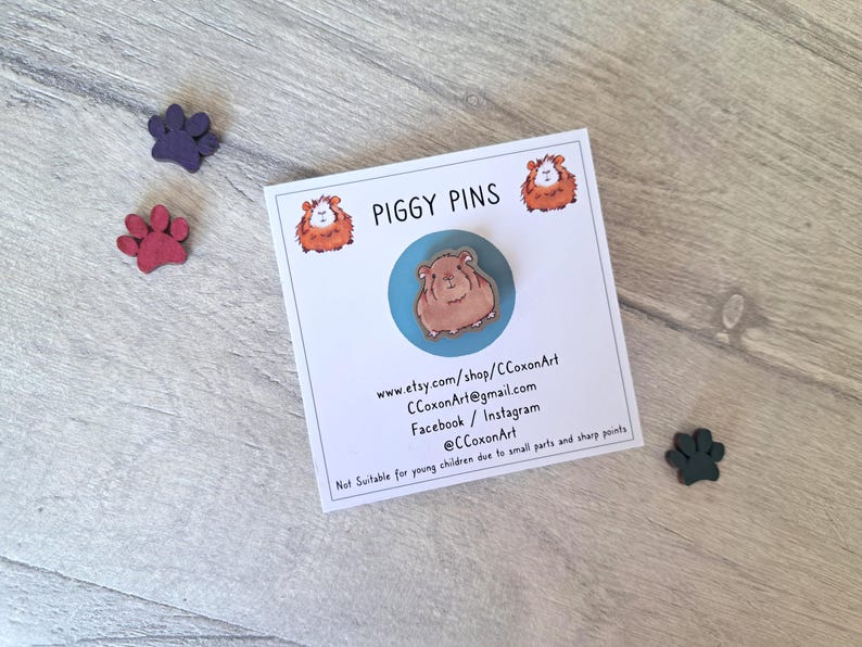 May include: A brown and white guinea pig pin on a blue background. The pin is part of a set of piggy pins. The card includes the shop name, website, and social media handles.