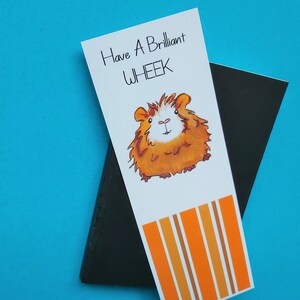 May include: A white bookmark with orange and brown stripes at the bottom. The bookmark features a cartoon illustration of a guinea pig with the text "Have a Brilliant Wheek" above the guinea pig.