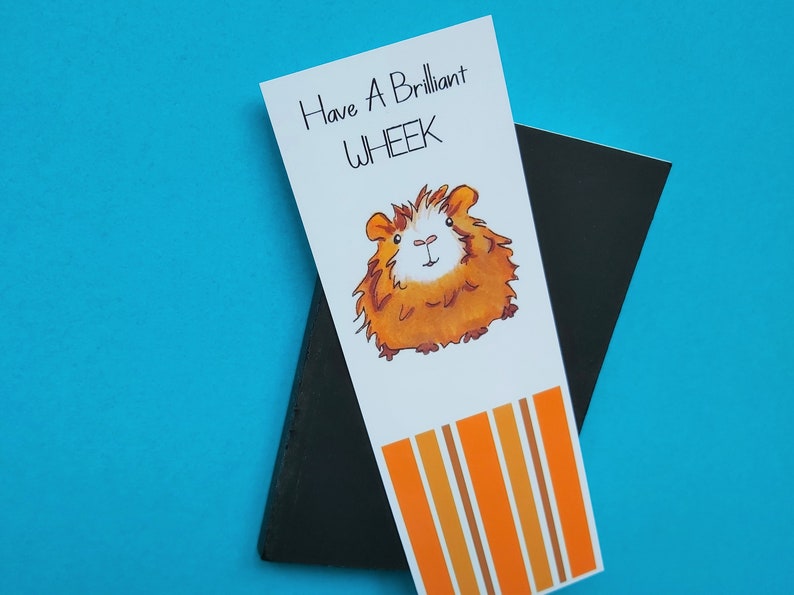 May include: A white bookmark with orange and brown stripes at the bottom. The bookmark features a cartoon illustration of a guinea pig with the text "Have a Brilliant Wheek" above the guinea pig.