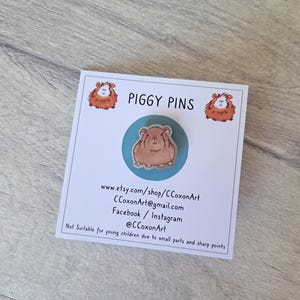 May include: A blue and white enamel pin featuring a brown and white guinea pig. The pin is on a white card with the text "PIGGY PINS" and contact information for the shop.
