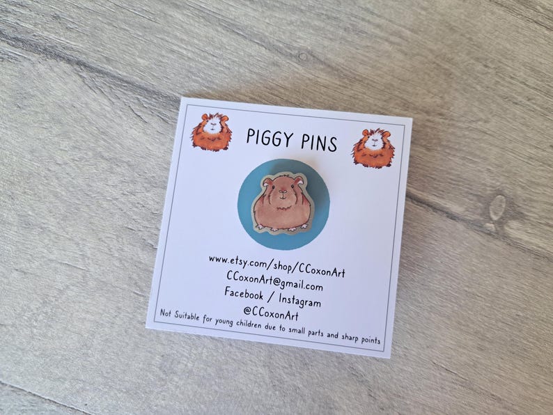 May include: A blue and white enamel pin featuring a brown and white guinea pig. The pin is on a white card with the text "PIGGY PINS" and contact information for the shop.