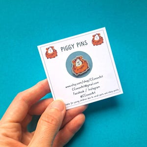 May include: A white card with the text "PIGGY PINS" and contact information for the shop. The card is held in a hand and features a small, round, enamel pin of a brown and orange guinea pig.