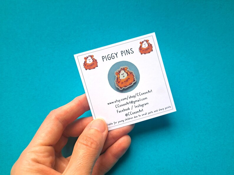 May include: A white card with the text "PIGGY PINS" and contact information for the shop. The card is held in a hand and features a small, round, enamel pin of a brown and orange guinea pig.