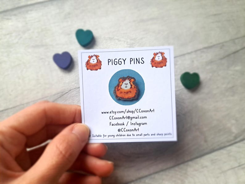 May include: A white card with the text "PIGGY PINS" and a cartoon image of a guinea pig. The card also includes contact information for the seller, including their Etsy shop, email address, and social media handles.