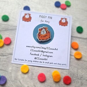 May include: A white card with the text "Piggy Pin for Amy" and a cartoon image of a guinea pig. The card also includes contact information for the artist, including their Etsy shop, email address, and social media handles.