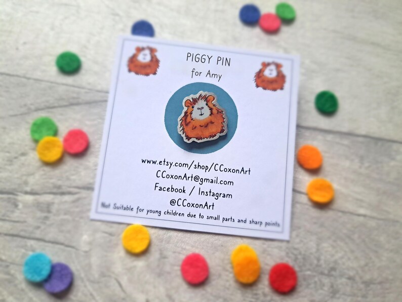 May include: A white card with the text "Piggy Pin for Amy" and a cartoon image of a guinea pig. The card also includes contact information for the artist, including their Etsy shop, email address, and social media handles.