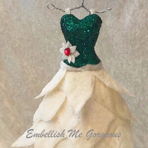 May include: A miniature dress ornament with a green sequined bodice and a layered, petal-like cream skirt. A white flower with a red center adorns the waist. The text "Embellish Me Gorgeous" is visible.
