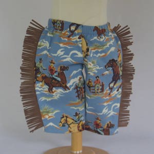 May include: Blue and white cowboy print children's pants with brown fringe trim. The pants are on a wooden mannequin.